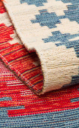 Close-up of the Ocean Form - Kilim - NMD04, a blue tribal rug with red, blue, and beige geometric patterns. One corner is folded to reveal the flatwoven texture and detailed stitching on the back.