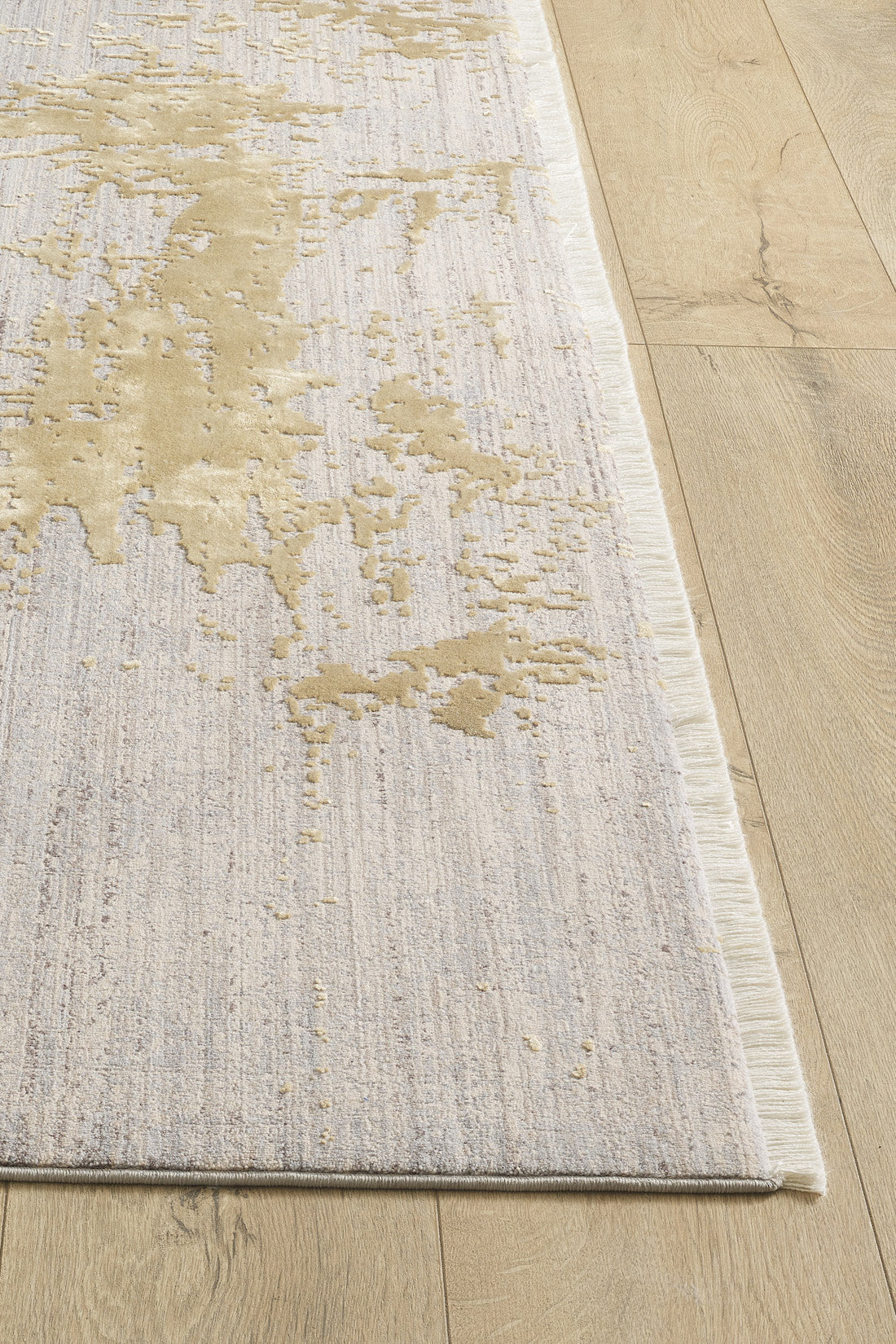 A close-up of the Old World Elegance Modern Rug - Yellow - M677O in beige, light gray, and yellowish gold abstract pattern on a light wood floor, featuring a subtle texture and thin fringe along the edge.