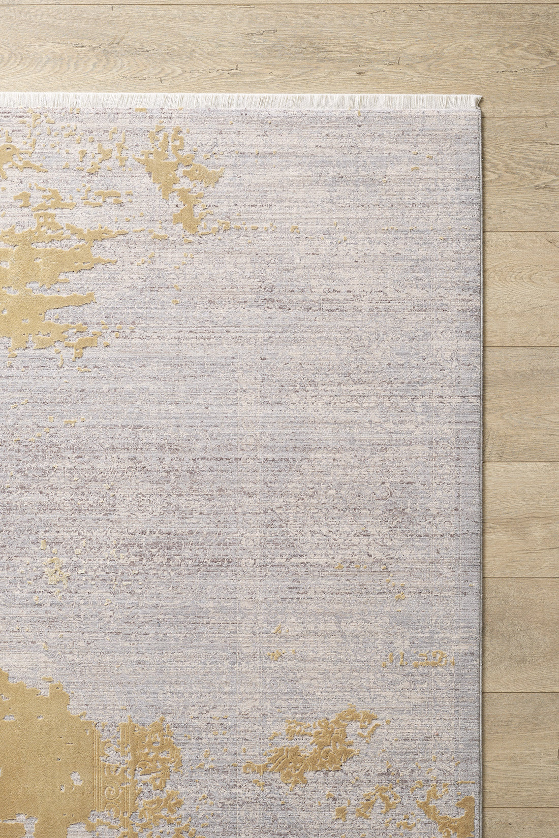 The Old World Elegance Modern Rug - Yellow - M677O, a machine-made viscose-polyester rug with a light grey, beige, and subtle gold abstract pattern plus fringe on the top edge, lies partially visible on a light wood floor.