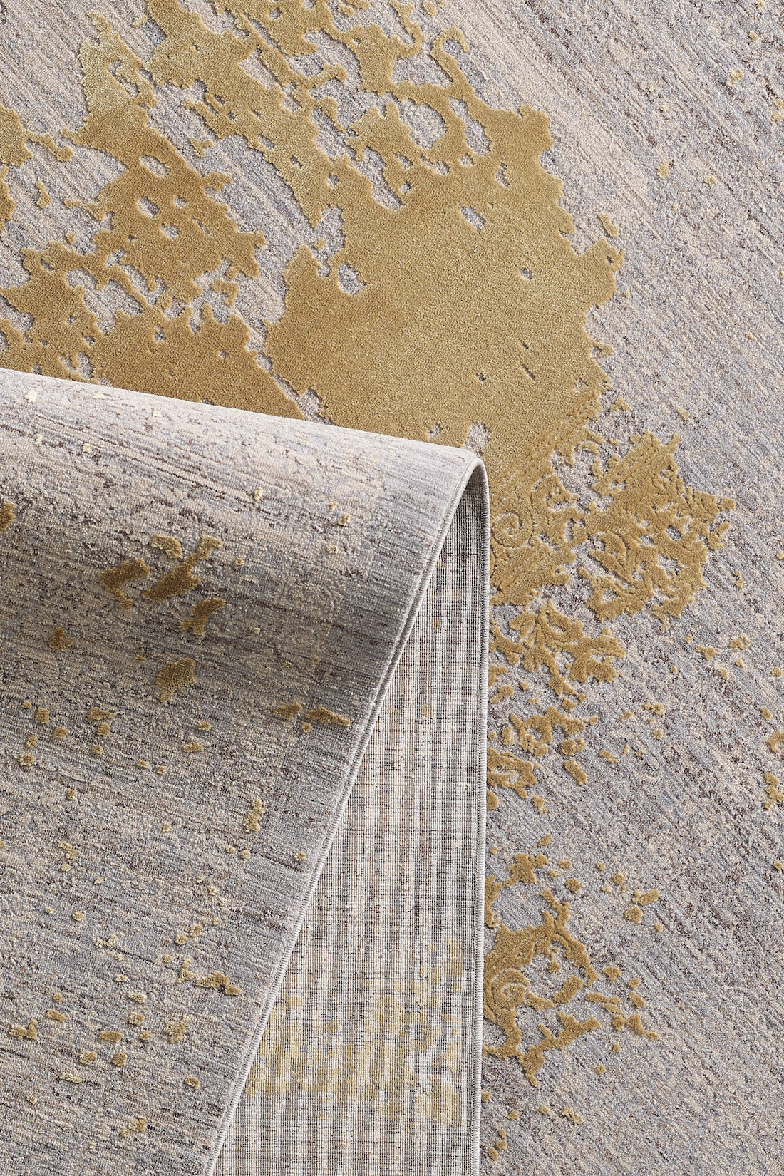 A close-up of the Old World Elegance Modern Rug - Yellow - M677O shows golden abstract patterns on a gray-beige background, with one corner folded to reveal the underside of this elegant machine-made rug.