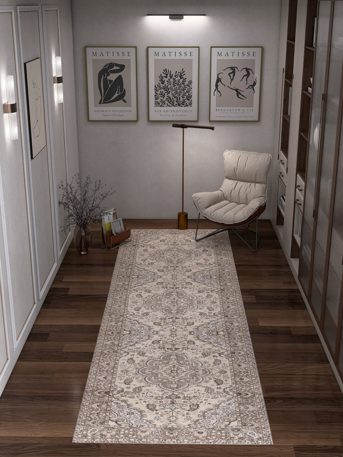 A hallway with dark wooden floors features the Opulent Mirage - Washable Rug - JRN1008 in a subtle medallion design, alongside a cushioned chair, floor lamp, glass vase with branches, framed Matisse prints, and shelves to the right.