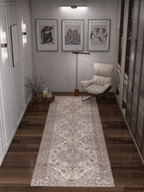 A hallway with dark wooden floors features the Opulent Mirage - Washable Rug - JRN1008 in a subtle medallion design, alongside a cushioned chair, floor lamp, glass vase with branches, framed Matisse prints, and shelves to the right.
