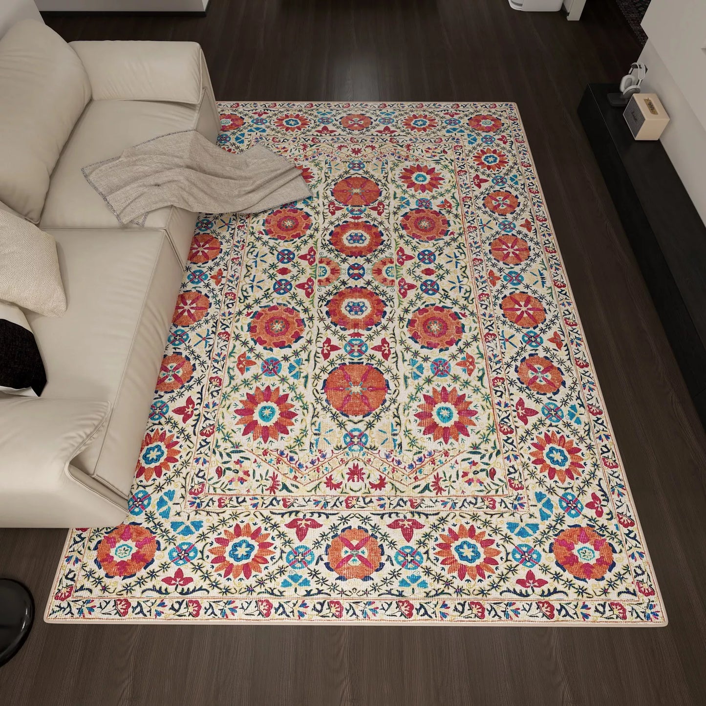 A living room with a cream sofa features the Orient Bloom - Washable Rug - JRN1029 (Custom Size), displaying red, blue, and green floral and geometric patterns on a light background, set atop dark wooden flooring.