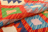 A close-up of the Oriental Harmony - Handwoven Kilim shows its colorful geometric patterns in red, green, blue, yellow, and orange. Folded layers reveal diamond shapes and zigzag designs that highlight the rug’s vibrant flatweave texture.