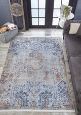 An Oriental Treasures - Machine-Washable Rug JR1613 (Custom Size) with blue, gold, and beige patterns is placed on a light wood floor by a gray sofa, checkered ottoman, and round table with a plant near glass doors.