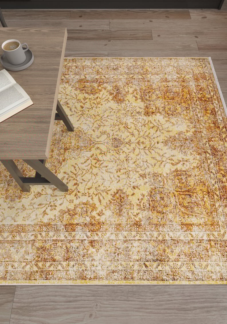 The Ornate Beauty - Washable Rug - JR206, featuring a rustic yellow-gold pattern, is displayed on a wooden floor beside a light wood table with coffee and an open book—ideal for high-traffic areas.
