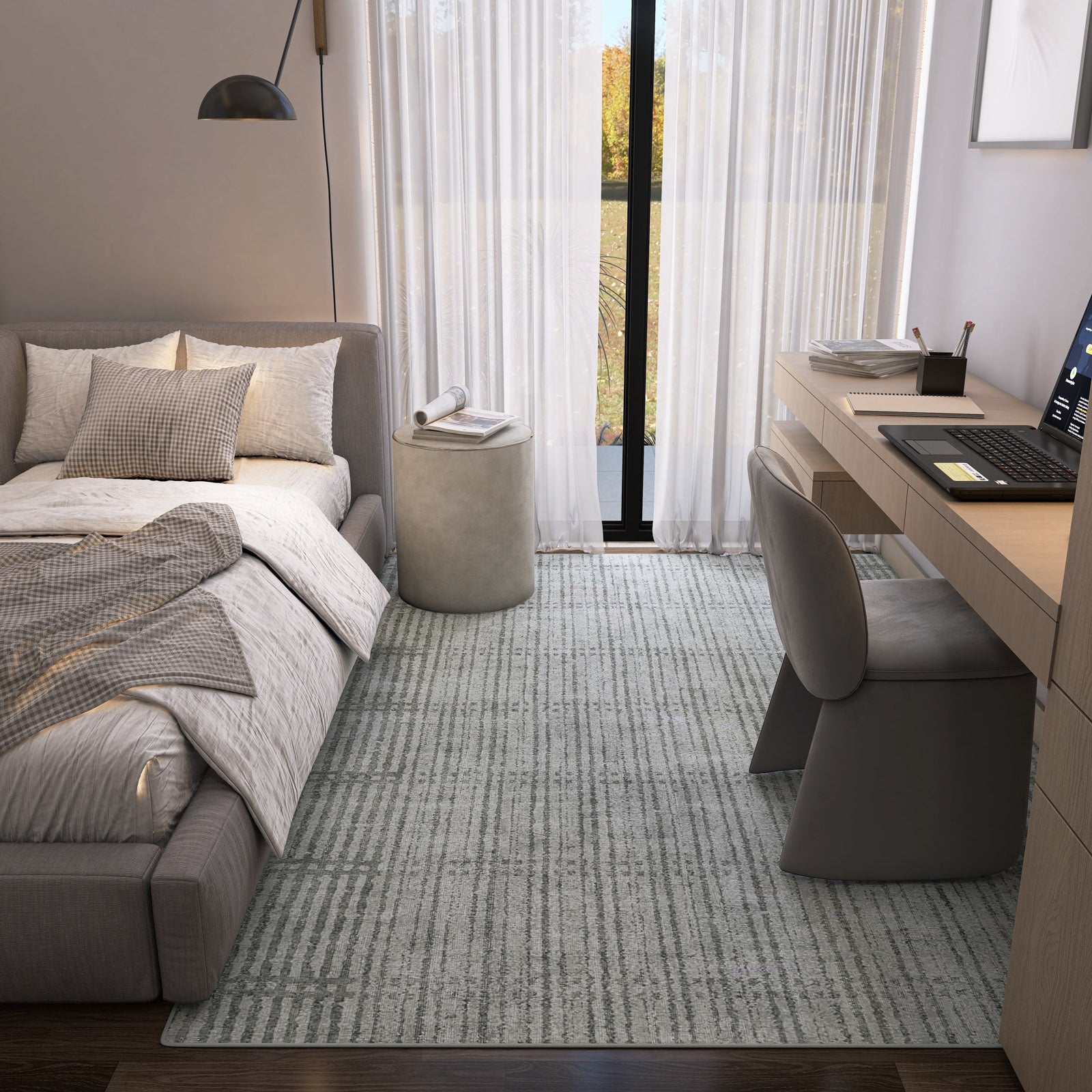 A modern bedroom features a gray upholstered bed, neutral bedding, an Ornate Hues - Washable Rug - JRN38 (Custom Size), a wooden desk with laptop and chair, a small round ottoman, and large windows with sheer curtains letting in natural light.