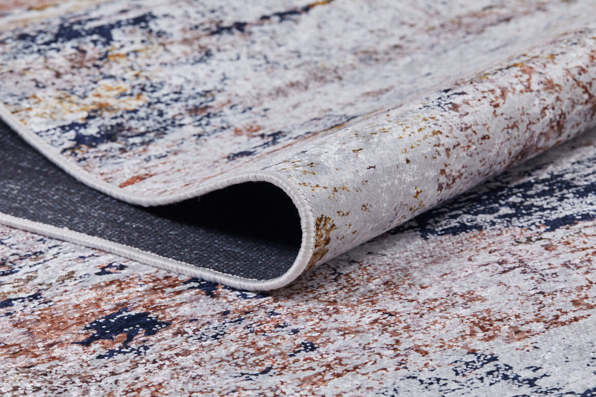A close-up of the Painted Dreams - Washable Rug - JR432 (Custom Size), featuring a textured abstract pattern in white, blue, brown, and gold with one edge curled to reveal the underside—perfect for allergy-sensitive and high-traffic areas.