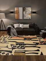 A modern living room with a black sofa, neutral cushions, a grey throw, two abstract wall artworks, a floor lamp, a small round side table with books, and the Pastel Harmony - Washable Rug - JRN43 featuring black and beige patterns.