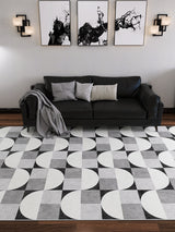 A modern living room with a black sofa, gray and white cushions, a gray throw blanket, abstract black-and-white wall art, and the Patina Bloom - Washable Rug - JRN50 with bold geometric circles and squares.
