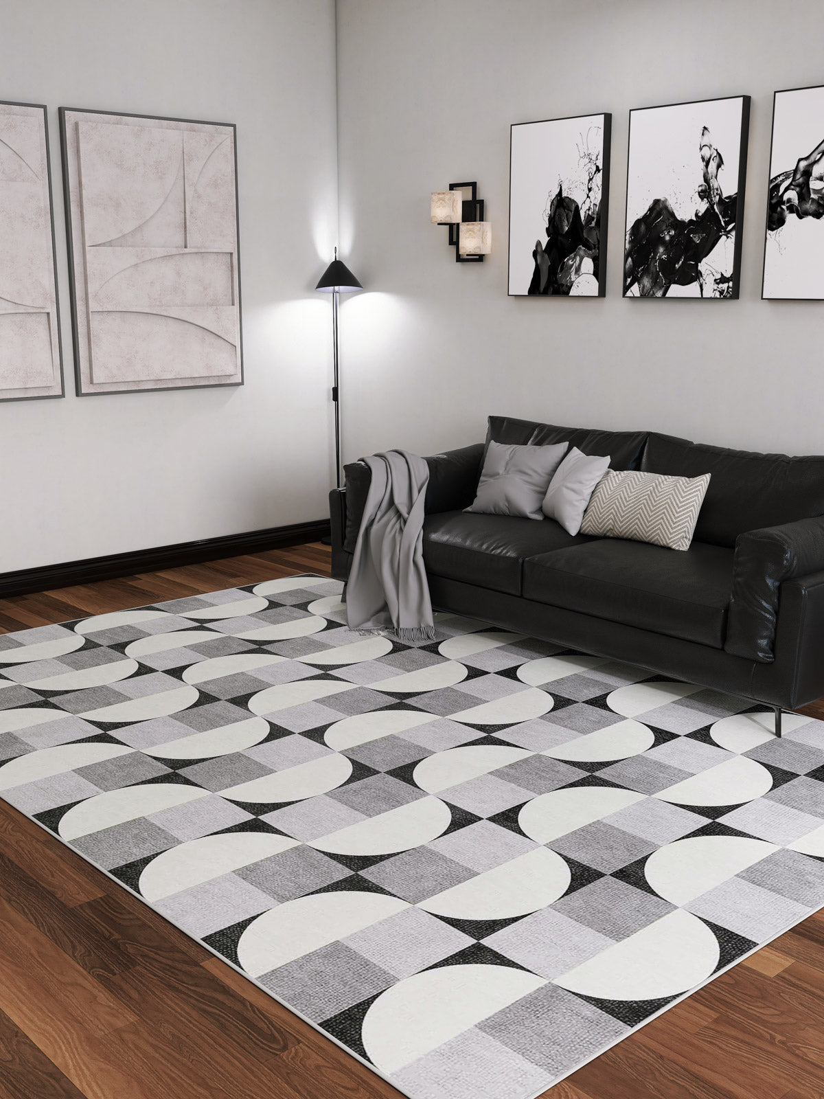 A modern living room with a black sofa, gray throw blanket, abstract wall art, floor lamp, wooden flooring, and the Patina Bloom - Washable Rug - JRN50. The space has a minimalist monochrome design with clean lines and contemporary decor.