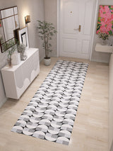 A bright, minimalist hallway with light wood floors features the Patina Bloom - Washable Rug - JRN50 as a geometric runner, paired with a white sideboard, potted plant, wall art, and a white door with gold hardware.