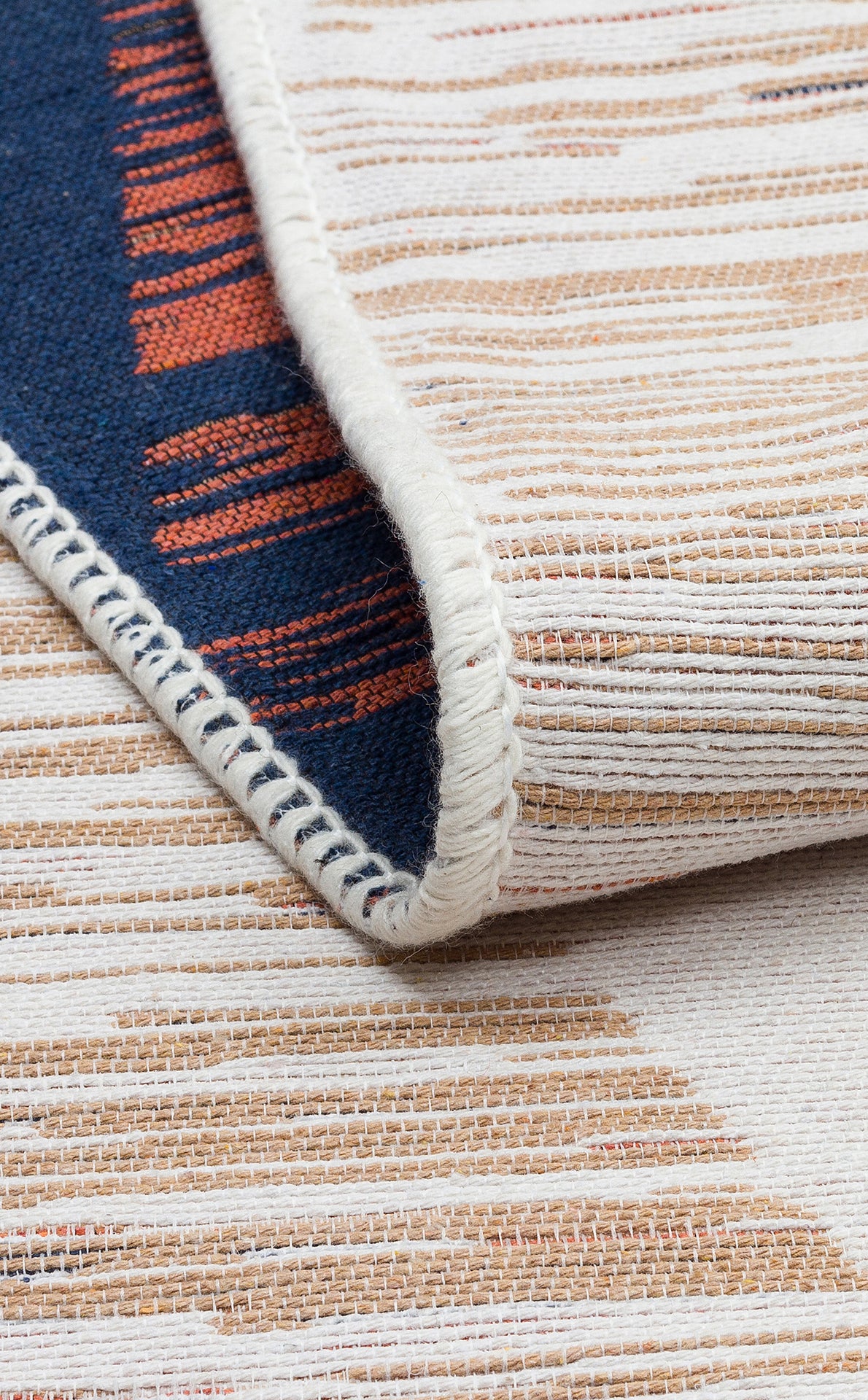 A close-up of the Pattern Field - Washable Reversible Kilims - AR38 flatwoven rug, showing beige, white, and blue threads with orange accent stripes and visible edge stitching. The partially folded rug reveals both reversible sides.
