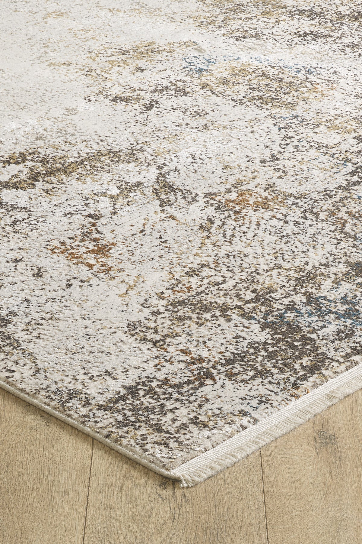 A close-up of the Pebble Pathway Premium Area Rug - M683L showcases its modern abstract, mottled pattern in neutral beige, gray, and brown tones on a light wood floor, with a visible machine-made edge and short fringe.