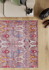A beige sofa with pillows sits beside the Peregrine Peachy Brown Washable Rug - LCC3022 (Outlet) US, featuring a colorful pattern on a wooden floor. Nearby are a stack of books, black binoculars, a black bag, and brown shoes.