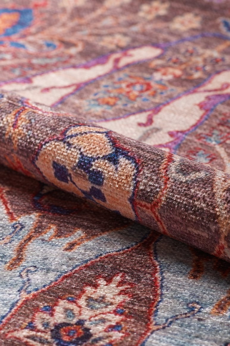 A close-up of a rolled corner of the Peregrine Peachy Brown Washable Rug - LCC3022 (Outlet) US shows detailed floral and geometric patterns in blue, red, beige, and brown on a flat weave surface.