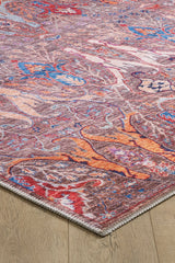 An angled close-up of the Peregrine Peachy Brown Washable Rug - LCC3022 shows its colorful abstract pattern in red, blue, orange, and purple on a light wood floor with detailed edge stitching.