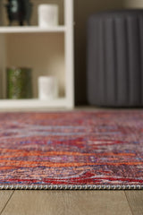 A close-up of the edge of the Peregrine Peachy Brown Washable Rug - LCC3022 (Outlet) US on a wooden floor, with a white shelving unit and gray ottoman softly blurred in the background.