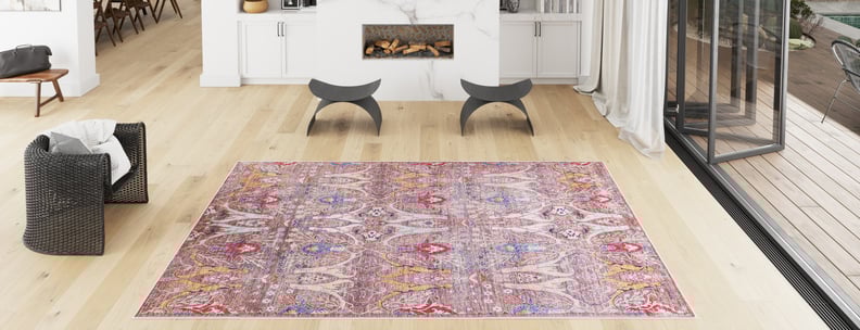 A spacious, modern living room with light wood floors features a Peregrine Peachy Brown Washable Rug (LCC3022), perfect for high-traffic areas, a black woven armchair, curved stools, a white marble fireplace, and floor-to-ceiling windows.