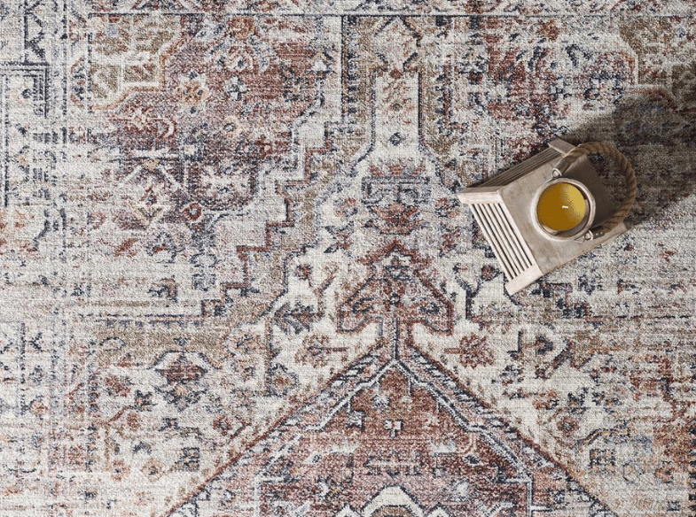 A patterned rug in earthy tones with a small tray holding a glass of orange juice placed in the upper right corner. Sunlight casts a shadow across part of the tray and drink.