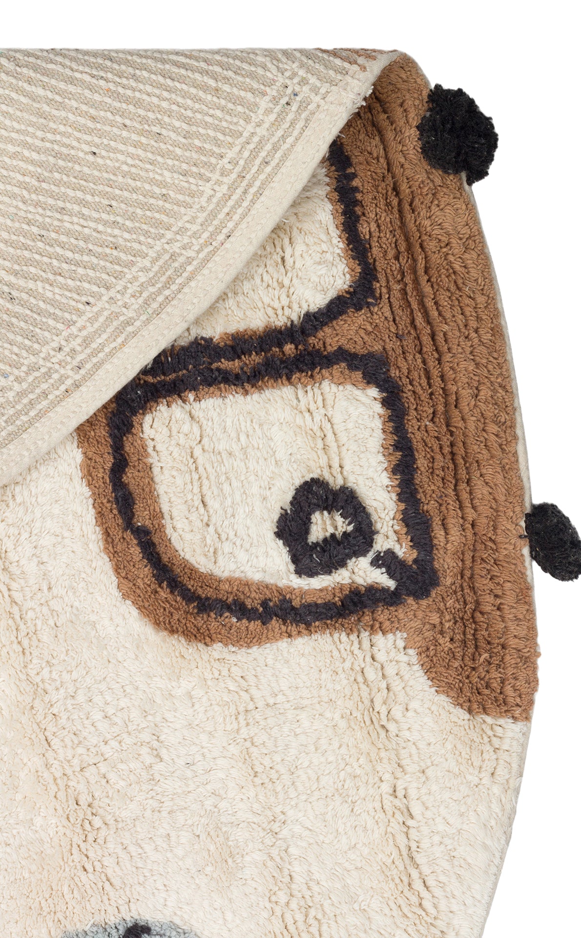 A close-up of the Play Patch - Washable Shaggy - CBN15 children’s rug, beige and brown with a cartoon face, black outlined eyes and glasses, two black pom-poms on the edge, and a corner folded to show its washable shaggy texture.