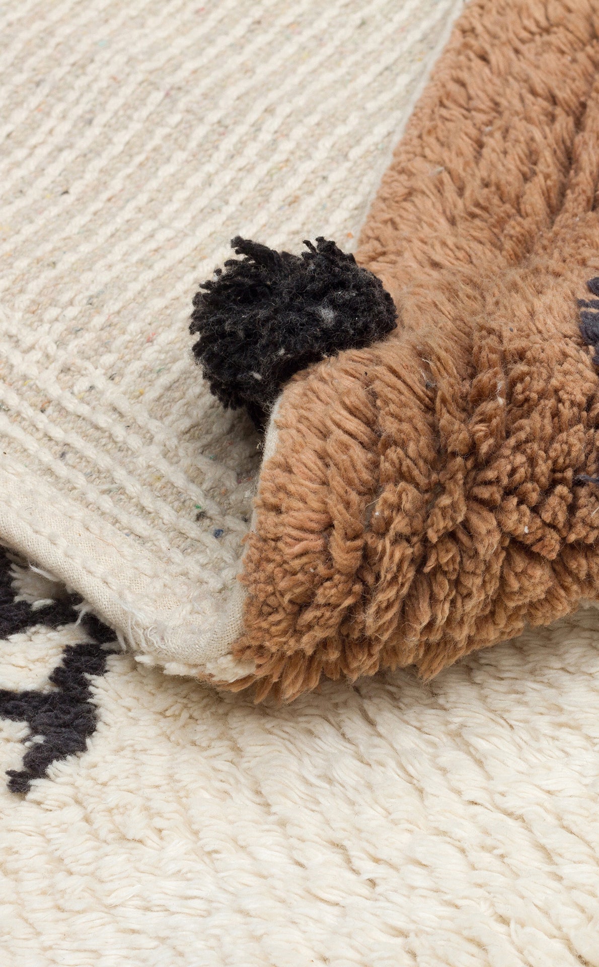 A close-up of the Play Patch - Washable Shaggy - CBN15 rug shows its brown fluffy top with a black tassel and pattern, folded to display the beige woven underside, resting on a cream-colored surface.