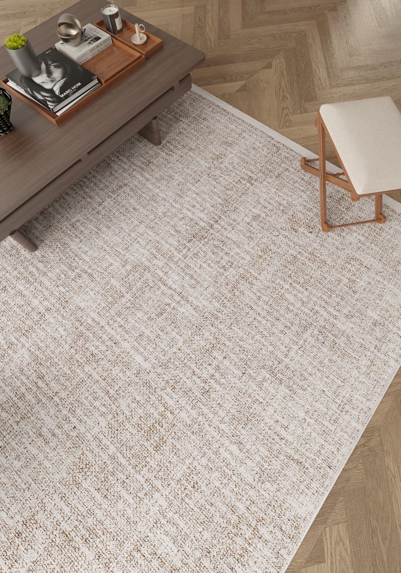 The Pure Bliss - Washable Rug - JR745, in light beige, sits on a herringbone wood floor beside a wooden coffee table with books, a tray, and a cup, plus a stool topped with a light cushion—ideal for high-traffic areas.