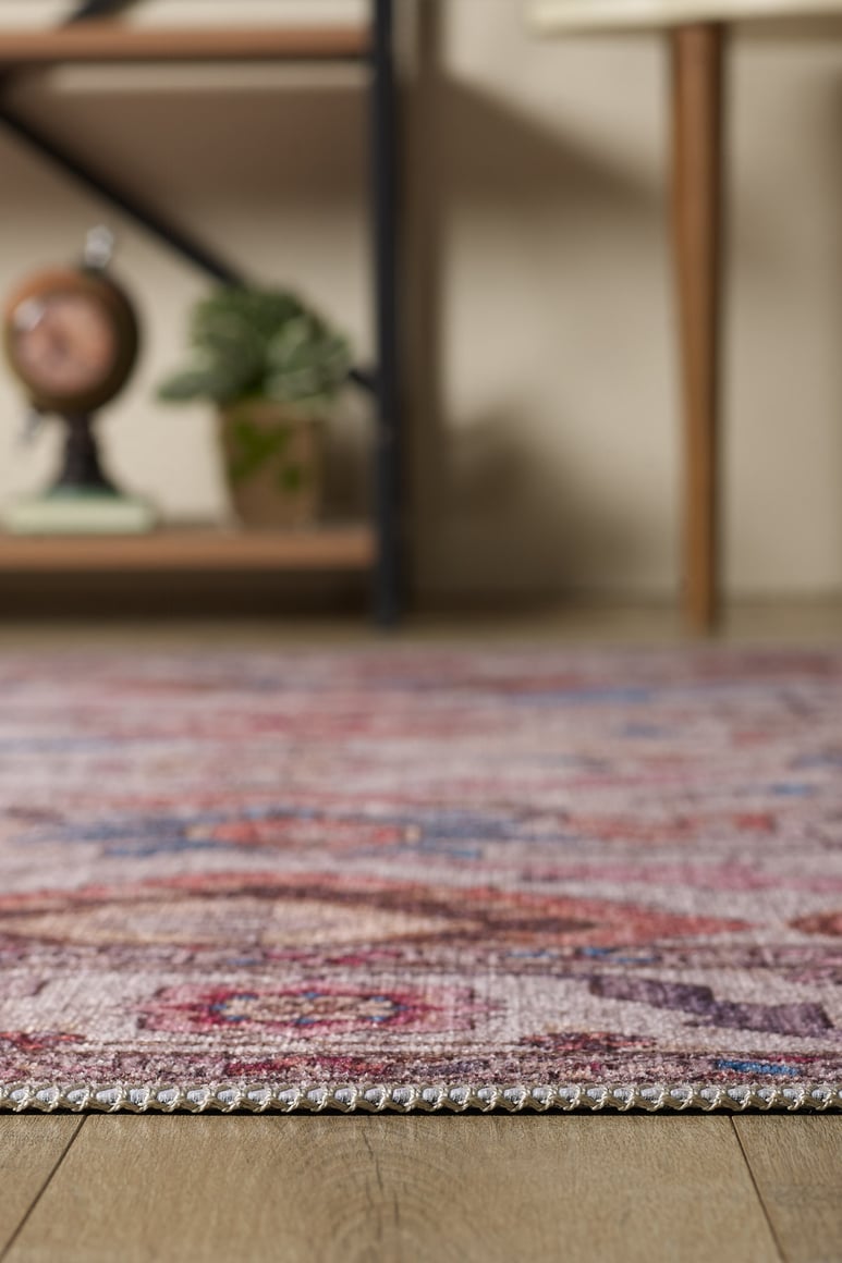 A close-up of the edge of the Quasar Traditional Washable Rug - LCC3024 in purple and pink on a wooden floor, with a blurred background showing a shelf, decorative clock, potted plant, and wooden furniture legs.