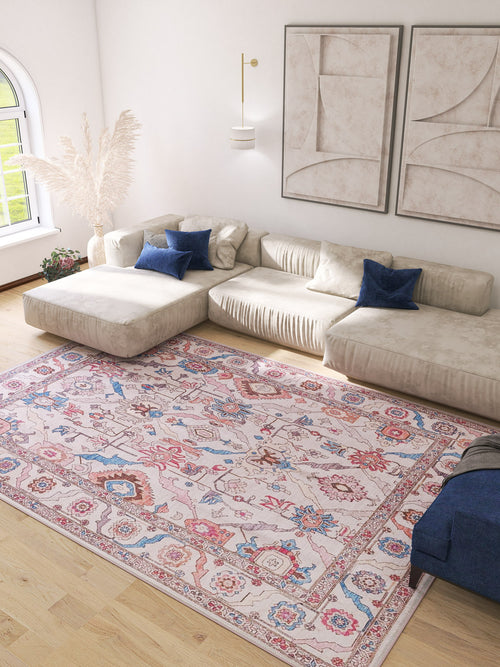 Load image 1 in gallery view. A bright living room with a cream sectional, blue accent pillows, abstract wall art, and the Quasar Traditional Washable Rug - LCC3024 in pink, blue, and red patterns on light wood floors. Sunlight pours in from an arched window.