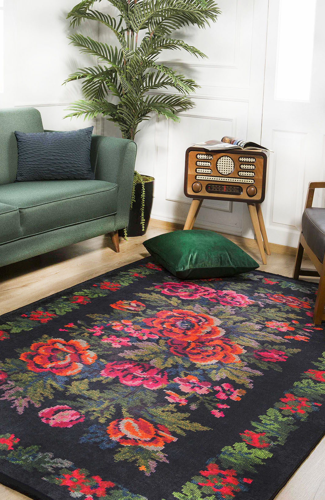 A living room features the Radiant Bloom - Washable Rug - JR294 (Custom Size) with bold red and pink florals on black, a green sofa, vintage radio on a wooden stand, potted plant, and a green velvet pillow on the floor.