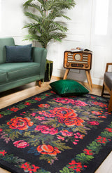 A living room features the Radiant Bloom - Washable Rug - JR294 (Custom Size) with bold red and pink florals on black, a green sofa, vintage radio on a wooden stand, potted plant, and a green velvet pillow on the floor.