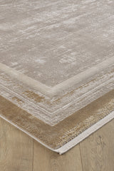 A close-up of the Radiant Edges Bordered Rug - Amber (M542G), a rectangular machine-made rug with a subtle beige and taupe pattern and layered neutral borders, displayed on a light wood floor.