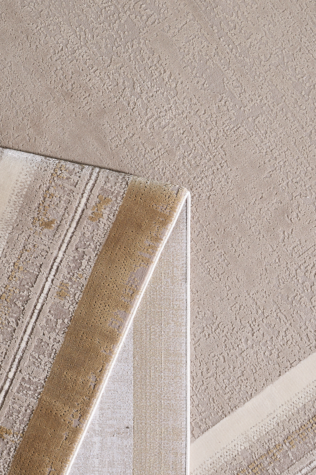 A close-up of a textured beige carpet with the Radiant Edges Bordered Rug - Amber - M542G folded at one corner, showing both its front and underside. The rug has neutral tones and geometric designs.