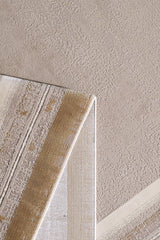 A close-up of a textured beige carpet with the Radiant Edges Bordered Rug - Amber - M542G folded at one corner, showing both its front and underside. The rug has neutral tones and geometric designs.