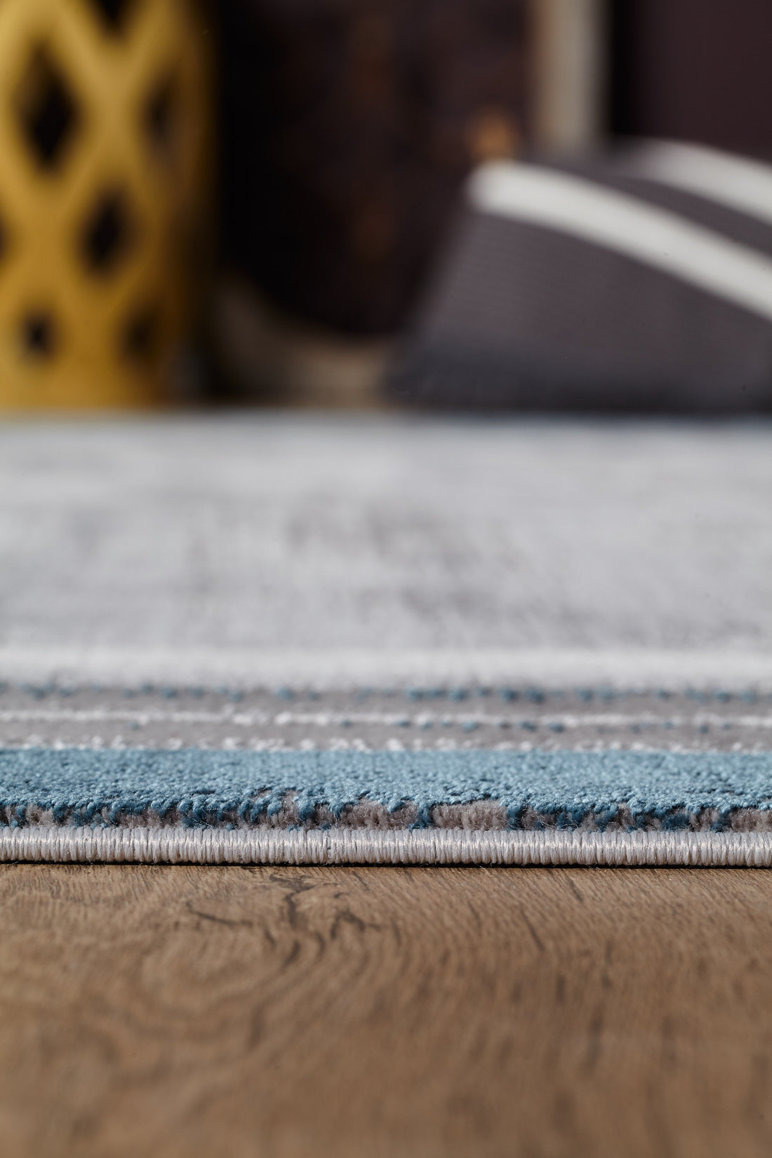 Close-up of the Radiant Edges Bordered Rug - Sapphire - M542I, showing its blue and gray edge design on a wooden floor, with blurred background items like a yellow decor piece and a striped cushion.