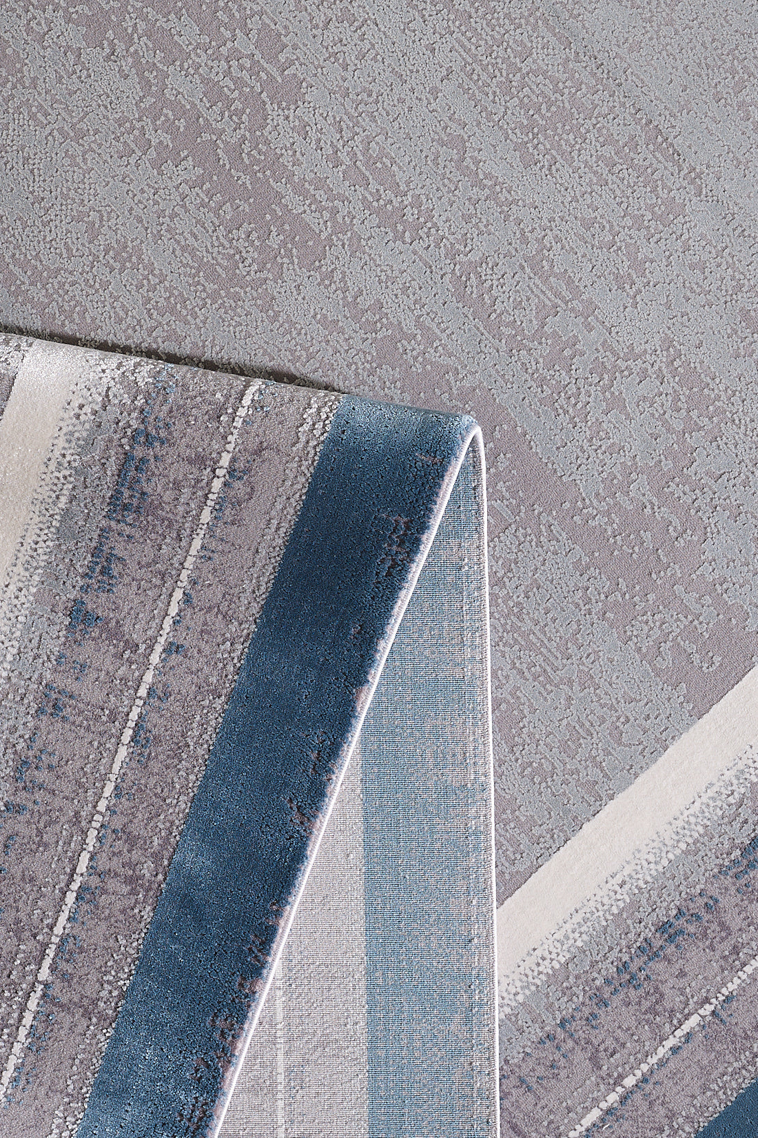 A close-up of the Radiant Edges Bordered Rug - Sapphire (M542I) shows a folded corner and a machine-made, textured surface with blue, gray, beige, and white stripes on a mottled gray background.
