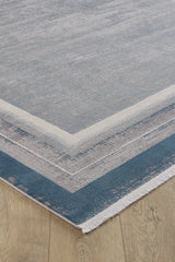 A close-up of the Radiant Edges Bordered Rug - Sapphire - M542I, a rectangular machine-made rug showing layered blue and gray tones, a gradient border design, and elegant white fringes on a light wooden floor.