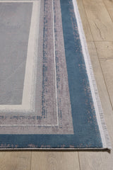 A close-up of the Radiant Edges Bordered Rug - Sapphire - M542I, a machine-made rug with blue, gray, and beige rectangular borders, subtle geometric pattern, and fringed edges on a light wooden floor.