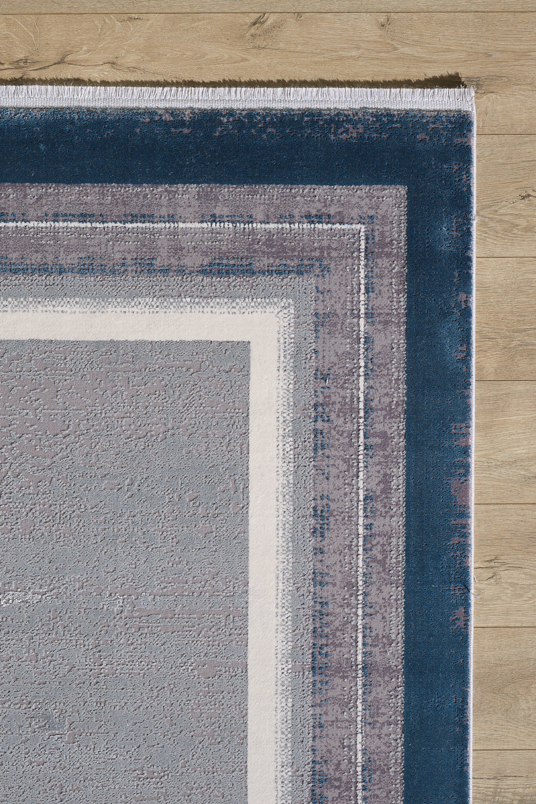 A close-up of the Radiant Edges Bordered Rug - Sapphire - M542I shows its rectangular layered blue, gray, and white borders and textured, slightly distressed machine-made look on a light wood floor.