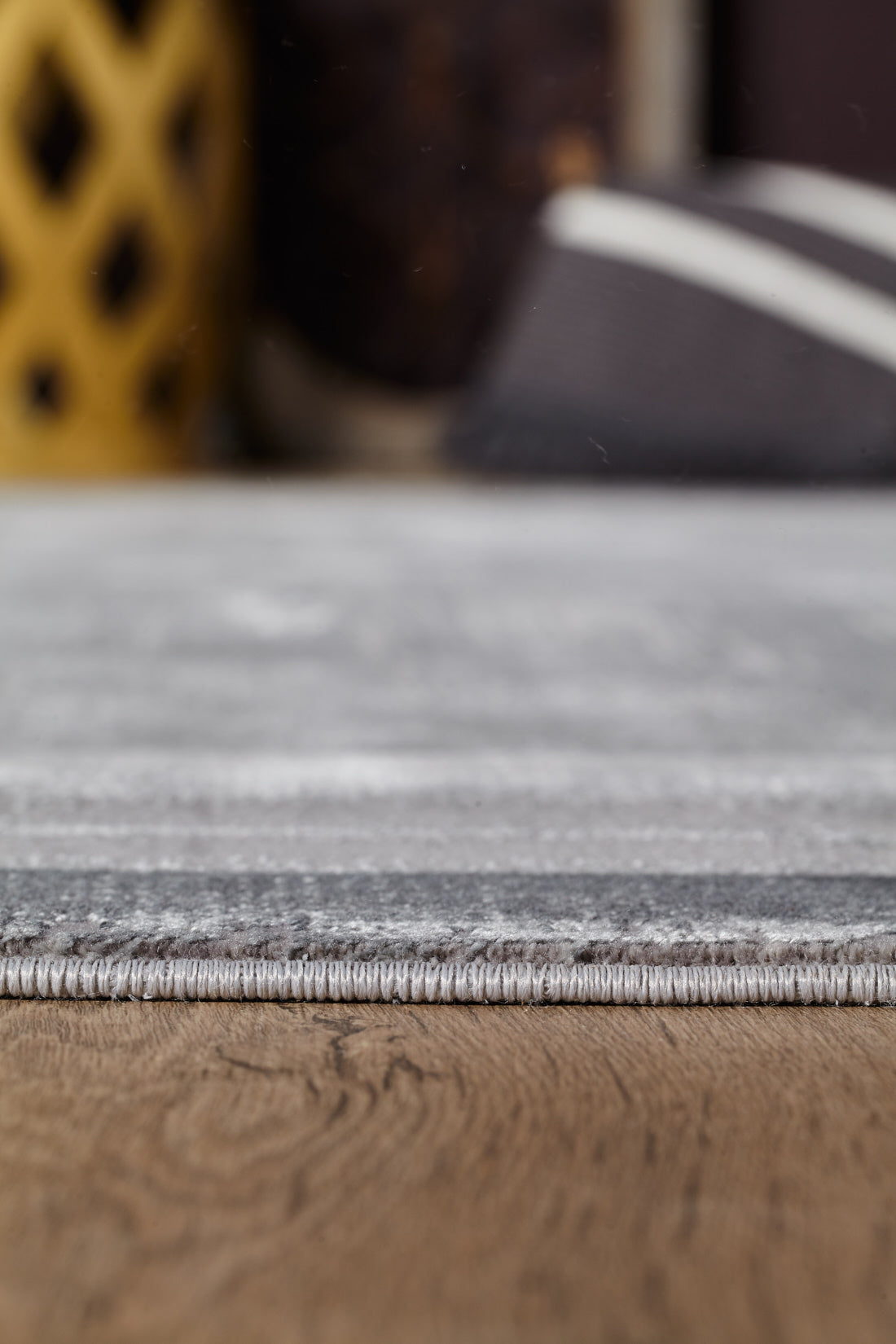 Close-up of the Radiant Edges Bordered Rug - Silver - M542B, featuring gray stripes and a stitched edge on a wooden floor. In the blurred background, a yellow decorative object and a black-and-white item are partially visible.