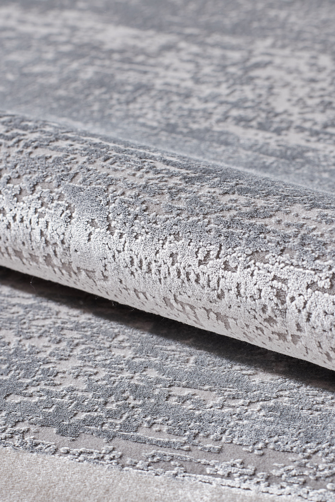 Close-up of the Radiant Edges Bordered Rug - Silver - M542B, showcasing its textured, metallic silver and gray surface with a shimmering pattern; rug is partially rolled to highlight its sheen and intricate design.