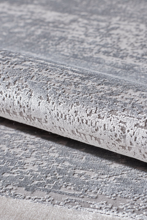 Load image 1 in gallery view. Close-up of the Radiant Edges Bordered Rug - Silver - M542B, showcasing its textured, metallic silver and gray surface with a shimmering pattern; rug is partially rolled to highlight its sheen and intricate design.