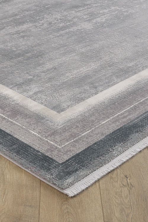 Load image 1 in gallery view. A close-up of the Radiant Edges Bordered Rug - Silver - M542B, a rectangular, flat-pile rug with a gray and light beige gradient pattern, short fringe on one edge, placed on a wooden floor. Machine-made for durability.