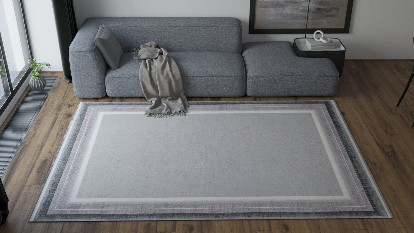 A modern living room with a gray sectional sofa, a light gray blanket on the arm, a rectangular coffee table, and the Radiant Edges Bordered Rug - Silver (M542B) featuring a chic border design on the wooden floor.
