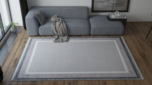 Load image 1 in gallery view. A modern living room with a gray sectional sofa, a light gray blanket on the arm, a rectangular coffee table, and the Radiant Edges Bordered Rug - Silver (M542B) featuring a chic border design on the wooden floor.
