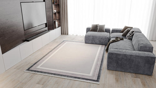 Load image 1 in gallery view. Modern living room featuring a gray sectional sofa, several cushions, built-in shelving, flat-screen TV, light wood flooring, white curtains, and the Radiant Edges Bordered Rug - Silver - M542B as the centerpiece.