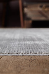 A close-up of the Radient Silver Haze Ombre Rug - M497D in light gray on a wooden floor, with a blurred background featuring part of a wooden chest or table.