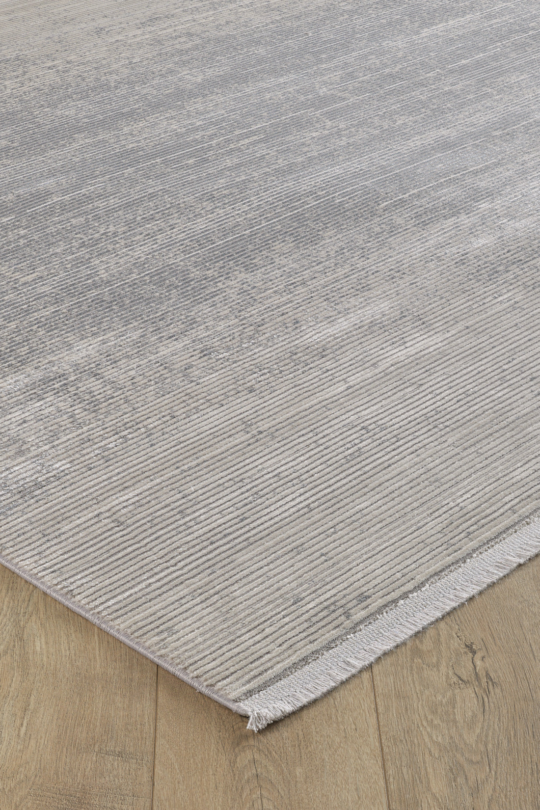A close-up of the Radient Silver Haze Ombre Rug - M497D, a minimalist rectangular light gray flat pile with subtle textured lines and short fringes, shown on a light wood floor. Machine-made for durability and style.