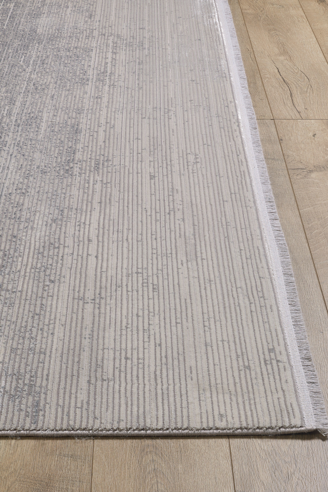 A close-up of the Radient Silver Haze Ombre Rug - M497D, featuring thin, uneven gray lines on a light background and short fringe on one edge, shown flat-pile style atop a wooden floor.