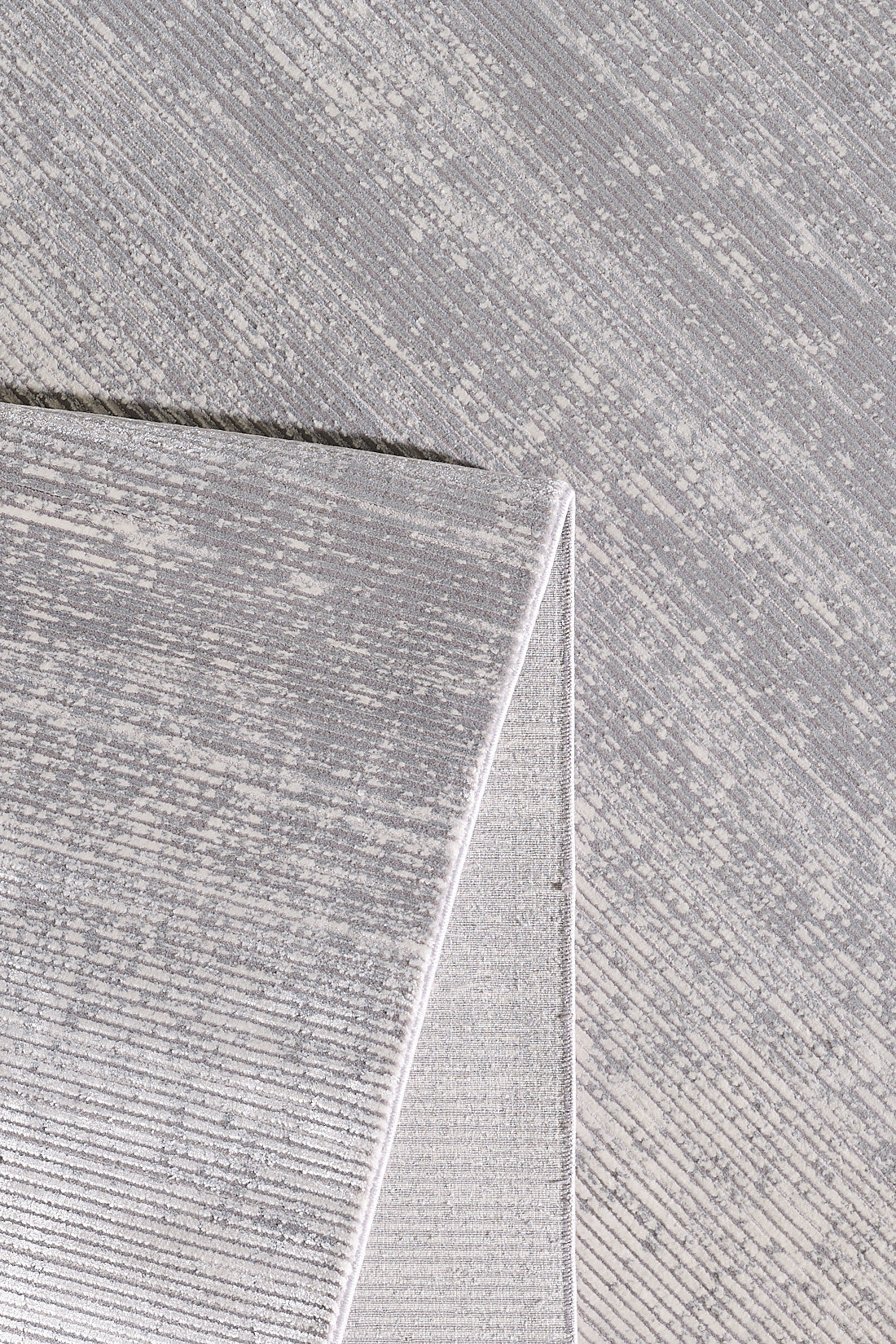 A close-up of the Radient Silver Haze Ombre Rug - M497D shows its textured light grey surface, a folded corner displaying the backing, and an intricate linear pattern on both sides.
